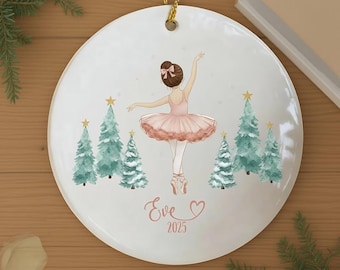 May include: A white ceramic Christmas bauble featuring a ballerina in a pink tutu and pointe shoes, surrounded by green Christmas trees. The bauble has a gold string for hanging and includes the name "Eve" and the year "2025".