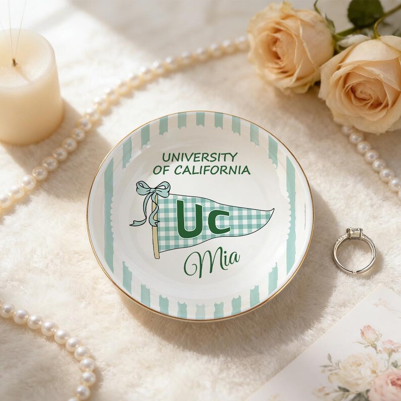 May include: A white ceramic ring dish with a gold rim, featuring a green and white striped pattern and a pennant design with the text "University of California" and "Uc Mia".