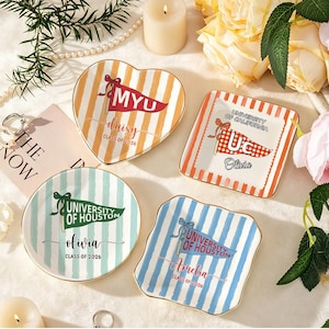 May include: Four decorative ceramic dishes with gold rims. Each dish features a different shape and design, including stripes and pennant flags with text. The text includes names, university names, and the year 2026.