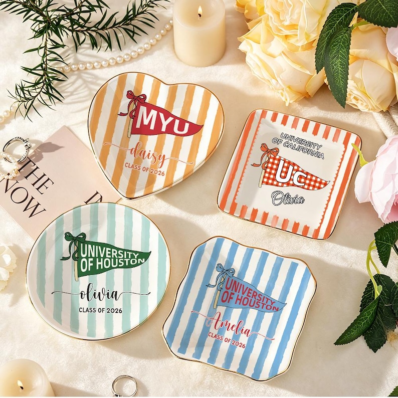 May include: Four decorative ceramic dishes with gold rims. Each dish features a different shape and design, including stripes and pennant flags with text. The text includes names, university names, and the year 2026.