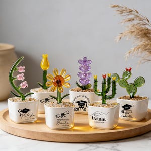 May include: A collection of glass flower sculptures in small white square pots, arranged on a wooden tray. The flowers include a sunflower, cactus, and lavender. Each pot has a graduation cap or scroll design with a name and "Class of 2020".