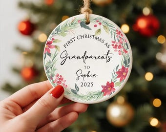 Peut inclure: Ornement de Noël rond en céramique avec l'inscription "First Christmas as Grandparents to Sophia 2025". L'ornement est orné d'une couronne florale rouge et verte, et possède une ficelle en jute pour l'accrocher.