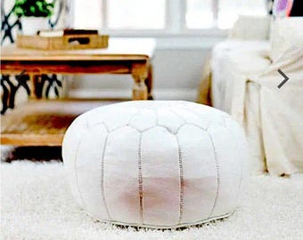 Op de afbeelding: Een witte, ronde Marokkaanse poef met een geometrisch patroon en verticale stiksels. De poef staat op een wit hoogpolig tapijt, wat duidt op een gezellige woonkamer. Het ontwerp is eenvoudig en elegant.