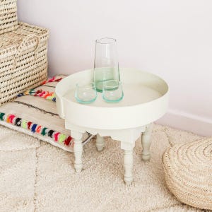 May include: A white, round side table with turned legs, holding a clear glass pitcher and two small, turquoise-tinted glasses. The table sits on a cream-colored rug, with a woven basket and a decorative pillow nearby.