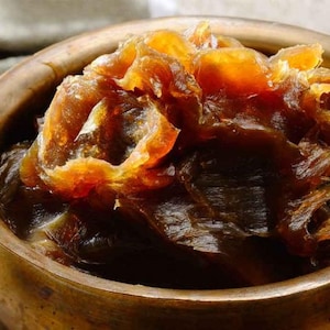 May include: Close-up of a wooden bowl filled with dark brown shea butter. The shea butter has a layered texture with golden-brown highlights. The bowl has a warm, natural wood grain appearance.