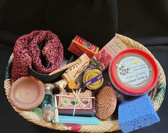May include: A woven basket filled with various spa products. Items include a black washcloth, a red loofah, a small clay bowl, a glass bottle, a comb, soap, and a jar of olive oil. The basket is on a black background.