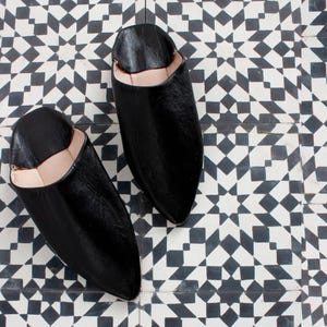 Puede incluir: Un par de zapatillas de cuero negro con punta y un interior de color claro. Las zapatillas se exhiben sobre un suelo de baldosas geométricas en blanco y negro. El calzado parece estar hecho a mano.