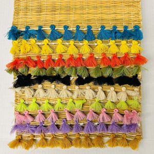 Könnte beinhalten: Eine gewebte Wanddekoration aus Stroh mit Reihen farbenfroher Quasten. Die Quasten sind in den Farbtönen Blau, Gelb, Orange, Grün, Schwarz, Creme, Limettengrün, Rosa, Lila und Gold gehalten. Die Quasten sind gleichmäßig angeordnet und erzeugen ein lebendiges, strukturiertes Design.