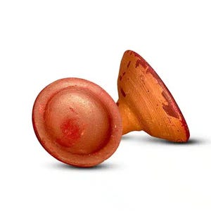 May include: Two handmade, terracotta-colored bottle stoppers. One stopper is round with a slightly raised center, and the other is a conical shape. Both have a reddish-brown hue with darker accents.