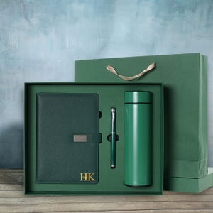 May include: A green gift set featuring a matching journal, pen, and insulated water bottle. The journal has a silver clasp and gold initials "HK". The set is presented in a green box with a matching gift bag.