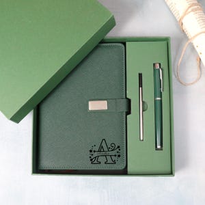 May include: A green gift set featuring a journal, pen, and refill in a matching box. The journal has a silver clasp and a custom initial design. The pen and journal are the same shade of green.