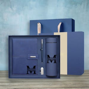 Puede incluir: Un juego de regalo azul que contiene un cuaderno, un bolígrafo, un vaso y una pequeña unidad USB. El cuaderno y el vaso están personalizados con el nombre "Marnie" y la letra "M". El juego se presenta en una caja de regalo azul con una bolsa a juego.