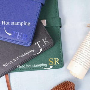 May include: Close-up of three personalized notebooks in blue, gray, and green. Each notebook features initials in hot stamping: blue with "TER", gray with "T.K", and green with "SR". The text "Hot stamping", "Silver hot stamping", and "Gold hot stamping" are also visible.