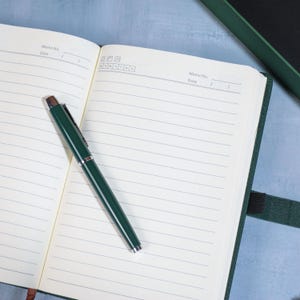 May include: An open, lined notebook with a dark green cover and a matching pen. The pen is resting on the open pages, which have a memo section at the top. The notebook is on a light blue surface.