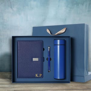 May include: A navy blue gift set featuring a notebook with a silver clasp, a pen, and a stainless steel water bottle. The notebook is embossed with the initials "K.P." The set is presented in a matching blue gift box and a blue gift bag.