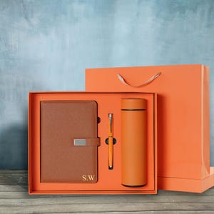 May include: An orange gift set featuring a brown leather notebook with the initials "S.W.", a matching orange pen, and a stainless steel water bottle. The set is presented in an orange box and gift bag.