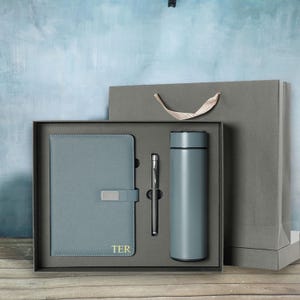 May include: A gift set in a gray box, featuring a gray notebook with a silver clasp and gold lettering, a silver pen, and a gray insulated water bottle. A gray gift bag is behind the box. The background is a textured blue wall.