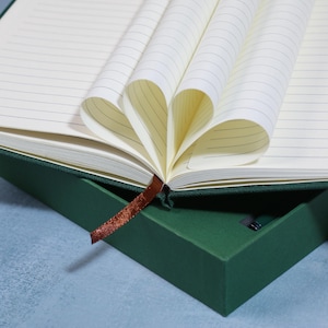 May include: A green journal with lined pages, a brown ribbon bookmark, and a matching green box. The pages are folded to create a heart shape. The journal is open, revealing the lined pages.