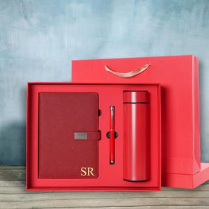 May include: A red gift set including a notebook with the initials "SR", a pen, and a thermos. The items are presented in a red box, with a red gift bag in the background. The notebook has a silver clasp.