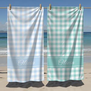 May include: Two beach towels with a gingham pattern, one light blue and one teal. Each towel has a white band with the name "Olivia" in cursive. The towels are hanging on a wooden rod with clothespins, with a beach and ocean background.