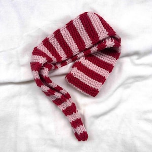 May include: A knitted hat and scarf set in a striped pattern of red and light pink. The hat is a long, pointed style, and the scarf is long and narrow, with a matching striped design. The set is made of a soft, textured yarn.