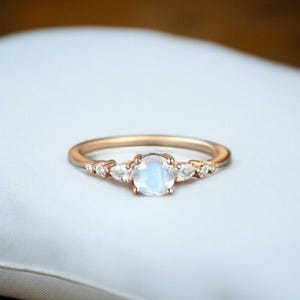 Puede incluir: Un anillo de oro rosa con una piedra central redonda de piedra lunar iridiscente, flanqueada por piedras de acento más pequeñas en forma de pera y redondas. El anillo está engastado en una banda delicada, mostrando un diseño clásico y elegante.