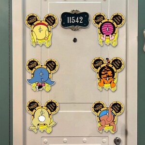 May include: A white door with a dark green trim features a house number plaque and six cartoon character magnets. The magnets are in the shape of Mickey Mouse ears, with yellow dripping accents and various character designs.