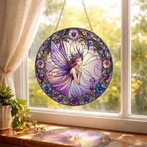 May include: A round stained glass window hanging from a gold chain. The artwork depicts a fairy with purple wings and a floral crown, surrounded by a wreath of flowers in shades of purple and blue. The window is set against a bright, sunlit background.