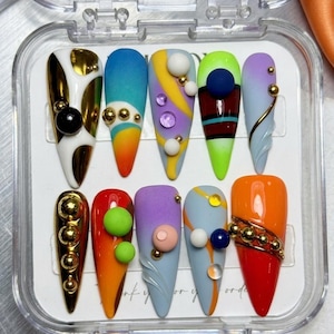 Könnte beinhalten: Ein Set aus zehn langen, spitz zulaufenden Kunstnägeln in einer transparenten Kunststoffbox. Jeder Nagel hat ein einzigartiges Design mit verschiedenen Farben, darunter Gold, Weiß, Blau, Orange und Lila. Verzierungen umfassen goldene Perlen, Kugeln und Wirbel.