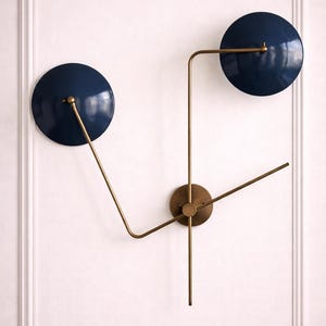 May include: A modern wall clock with a minimalist design. It features two navy blue circular elements and brass-colored arms and a central hub. The clock is mounted on a white wall, creating a striking contrast.