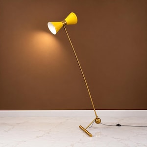 May include: A floor lamp featuring a bright yellow, double-cone shade with a white interior. The lamp has a long, thin, gold-toned metal stem connected to a spherical joint and a horizontal base bar. The light is illuminated, casting a warm glow.