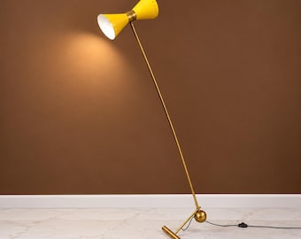 May include: A floor lamp featuring a bright yellow, double-cone shade with a white interior. The lamp has a long, thin, gold-toned metal stem connected to a spherical joint and a horizontal base bar. The light is illuminated, casting a warm glow.