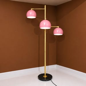 May include: A floor lamp with a tall gold-toned central pole and a round black base. The lamp features three adjustable arms extending from the pole, each holding a dome-shaped pink shade with a white interior, providing directional lighting.