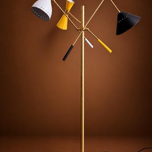 May include: A floor lamp with a central brass pole and a round black base. It features three adjustable arms with conical shades in white, yellow, and black. The lamp includes three smaller decorative rods in black, white, and yellow accents.
