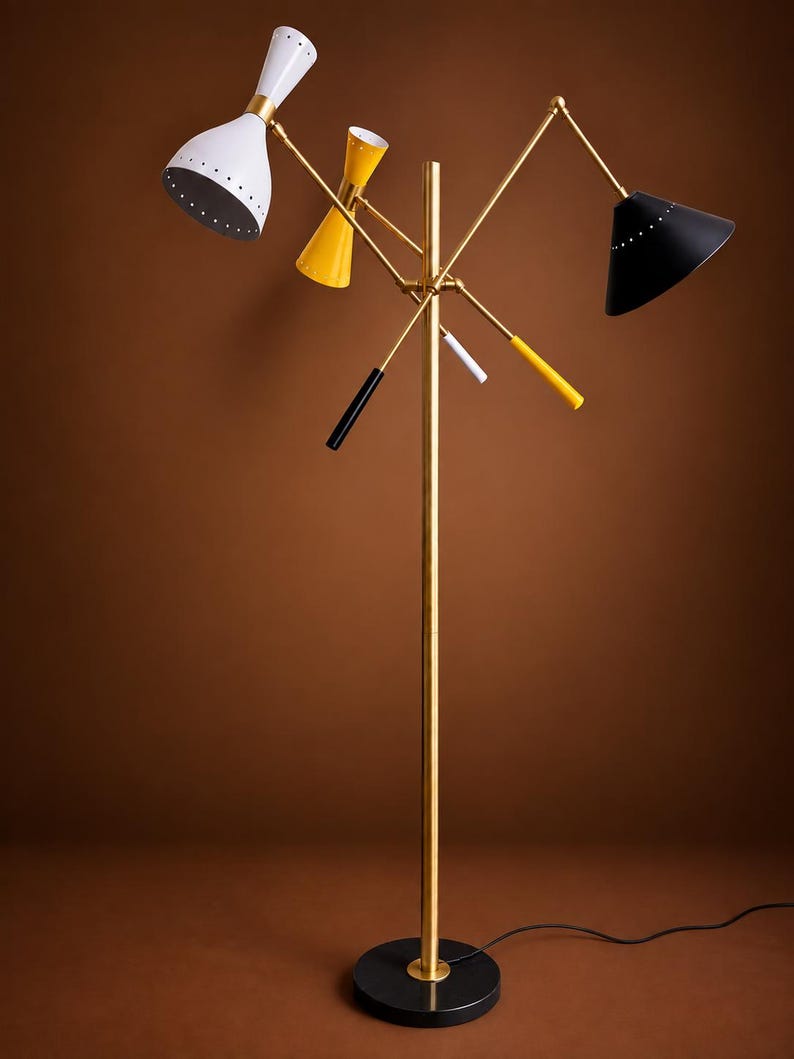 May include: A floor lamp with a central brass pole and a round black base. It features three adjustable arms with conical shades in white, yellow, and black. The lamp includes three smaller decorative rods in black, white, and yellow accents.