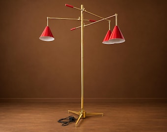May include: A floor lamp with a central brass pole and a tripod base. It features three adjustable arms with red conical shades and white interiors. The arms have red handles for positioning, and a black power cord extends from the base.