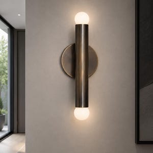 May include: A wall-mounted light fixture featuring a vertical, cylindrical dark bronze metal body attached to a circular backplate. Two small, spherical white glass bulbs are positioned at the top and bottom ends, emitting a soft, warm glow.