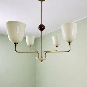 May include: A mid-century modern chandelier with four off-white, tulip-shaped shades. The fixture has brass arms and a dark brown sphere. The ceiling mount is brass, and the fixture hangs in a room with pale green walls.