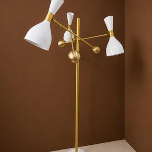 May include: A floor lamp featuring a central gold-toned metal pole mounted on a circular white marble base. The lamp has three adjustable arms with white conical shades and gold accents, complemented by decorative gold spheres on the central stem.