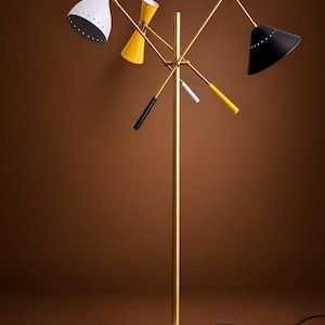 May include: A floor lamp with a brass central pole and a white marble circular base. It features three adjustable arms with conical shades in white, yellow, and black, each with small perforations along the rim and matching colored handles.