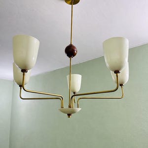 May include: A mid-century modern chandelier with a brass finish and five off-white glass shades. The fixture has a central wooden sphere and a circular base. The light hangs from a brass ceiling mount.