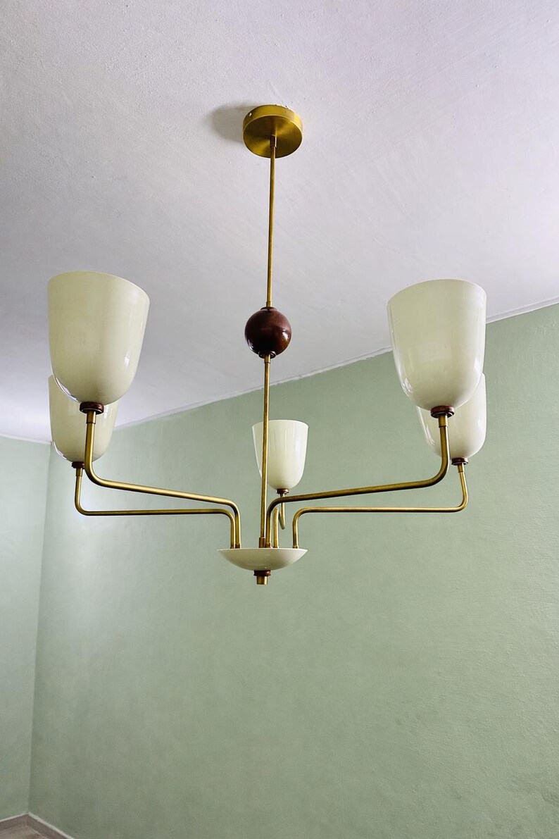May include: A mid-century modern chandelier with a brass finish and five off-white glass shades. The fixture has a central wooden sphere and a circular base. The light hangs from a brass ceiling mount.