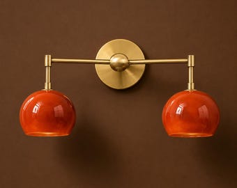May include: A wall-mounted light fixture featuring a horizontal brass arm attached to a circular brass backplate. Two downward-facing, glossy orange glass globes are suspended from the ends of the arm, creating a symmetrical lighting design.