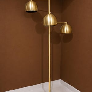 May include: A floor lamp with a tall, slender brass-colored central pole and a circular black base. The lamp features three dome-shaped brass-colored shades attached to adjustable arms at varying heights, directing light downward from the fixture.