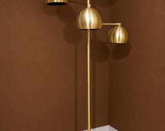May include: A floor lamp with a tall, slender brass-colored central pole and a circular black base. The lamp features three dome-shaped brass-colored shades attached to adjustable arms at varying heights, directing light downward from the fixture.
