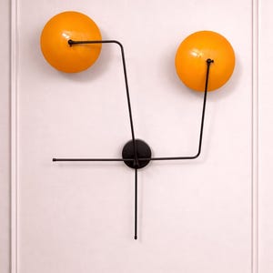 May include: Modern wall sconce featuring two spherical, orange-yellow light fixtures connected by black metal arms to a central black disc. The design is mounted on a white wall with decorative molding, creating a minimalist aesthetic.