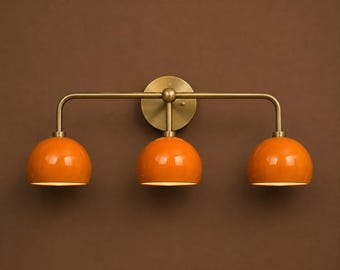 May include: A three-light wall fixture featuring a horizontal brass bar mounted to a circular brass wall plate. Three glossy orange dome-shaped shades are attached to the bar, each pointing downward to provide focused illumination from the light source.