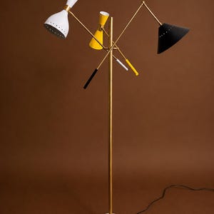 May include: A floor lamp with a central brass pole and a round white marble base. It features three adjustable arms with conical shades in white, yellow, and black, each with small perforations along the rim. The lamp includes thin brass rods.