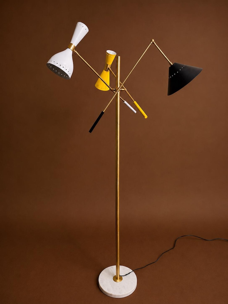 May include: A floor lamp with a central brass pole and a round white marble base. It features three adjustable arms with conical shades in white, yellow, and black, each with small perforations along the rim. The lamp includes thin brass rods.