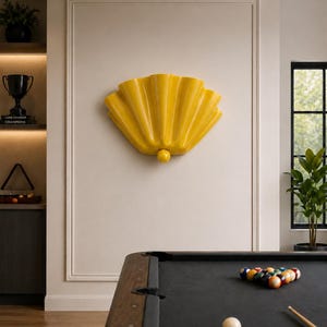 May include: A yellow, fan-shaped wall sconce with a rounded base is mounted on a white wall. A pool table with a black felt surface and a rack of billiard balls is in the foreground. A trophy and a plant are visible in the background.